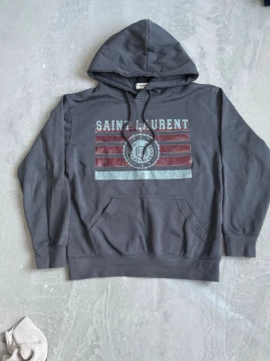 Mörkgrå Saint Laurent hoodie - Mörkgrå hoodie från Saint Laurent med tryck i rött och vitt framtill. Klassisk huva med dragsko och stor magficka. Snygg streetstil med grafiskt mönster och text. Perfekt för dig som gillar en avslappnad look.