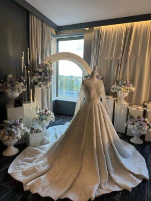 Glittrig lång brudklänning med släp - Magisk långklänning i glittrande vitt tyg med v-ringning och långa ärmar. Klänningen har ett dramatiskt släp och eleganta spetsdetaljer på ärmarna. Perfekt för dig som vill sticka ut och känna dig som en prinsessa.