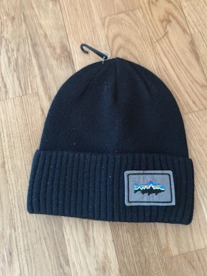 Svart mössa från Patagonia - Svart ribbstickad mössa från Patagonia med uppvikt kant och en tygpatch med logga framtill. Klassisk modell som passar perfekt till kalla dagar. Materialet känns mjukt och värmande.