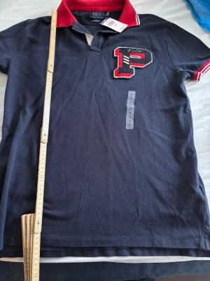 Svart Polo Ralph Lauren piké - Svart pikétröja från Polo Ralph Lauren med röd krage och vita/röda ränder på ärmslut. Stor broderad P på bröstet och texten 'POLO NEW YORK' på ryggen. Snygg kontrast mellan svart och rött, klassisk college-vibe. Perfekt för dig som gillar streetwear och sportig stil , 65cm längd , 50cm bred