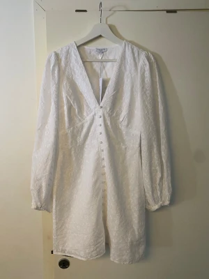 Vit kort klänning från bubbleroom  - Supersöt vit kort klänning från bubbleroom med v-ringning, långa ärmar och knappar framtill. Klänningen har broderade blommor i tyget och är gjord i bomull. Den är aldrig använd! nypris 699