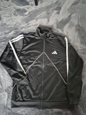 Svart Adidas träningsjacka - Snygg svart träningsjacka från Adidas med vita ränder längs ärmarna och dragkedja framtill. Jackan har hög krage, muddar vid ärmslut och nederkant samt Adidas-logga på bröstet. Tillverkad i ett glansigt, sportigt material som passar perfekt till träning.