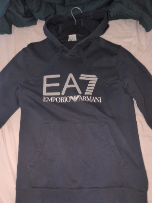 Mörkblå EA7 Emporio Armani hoodie - Snygg mörkblå hoodie från EA7 Emporio Armani med stor logga framtill. Klassisk känguruficka och justerbar huva med dragsko. Perfekt för en chill och stilren look. Mjuk bomullskänsla och långa ärmar. Säljer pga för liten