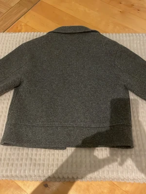 Grå ullkappa från Lager 157, kort modell - Stilren grå kappa från 157 i kortare modell. Kappan har klassisk krage och är tillverkad i ett ull-liknande material som passar perfekt för kyligare dagar. Enkel och tidlös design som funkar till många olika outfits.