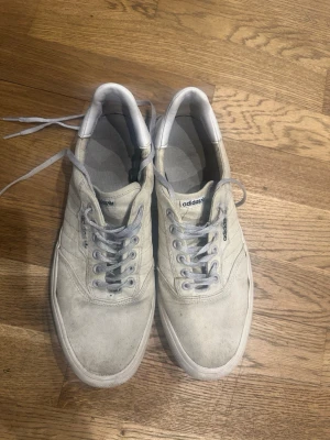 Vita Adidas sneakers i mocka - Säljer ett par vita Adidas sneakers i mocka med klassisk låg profil och snörning. Skorna har diskreta ränder på sidan och Adidas-logga på plösen. Perfekta för en clean och avslappnad stil.