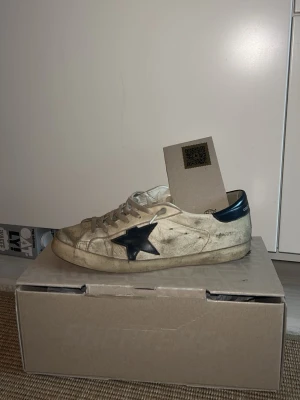 Golden Goose vita sneakers med svart stjärna - ÄKTA GOLDEN GOOSE sneakers i vitt läder med marinblå stjärna. Lite använda därav lägre pris. Perfekt för den som vill ha ett par exklusiva skor till våren utan att behöva pröjsa för mycket. ÄKTA kan du kolla genom att scanna skon där fram. 