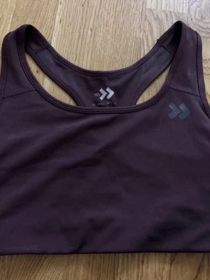 Mörklila sport-bh från Dobsom - Snygg mörklila sport-bh från lager med brottarrygg och disk Benchmark-loggaoty på bröstet. Tillverkad i soprtigt, stretchigt material som andas och passar perfekt till träning eller gym. Tight passform och mjuk känsla.