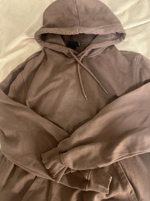 Brun hoodie med huva och magficka - Säljer en enkel brun hoodie med huva och dragsnören. Hoodien har en stor magficka framtill och ribbade muddar vid ärmslut och nederkant. Tillverkad i mjukt bomullsliknande material, perfekt för en avslappnad stil.