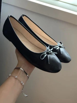 Svarta ballerinaskor med rosett - Stilrena svarta ballerinaskor med rund tå och söt liten rosett framtill. Skorna har en slät ovandel i ett matt material och ljus innersula. 