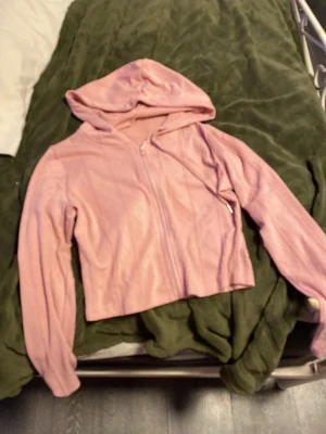 Rosa croppad hoodie med dragkedja - Säljer en ljusrosa croppad hoodie med dragkedja framtill och huva. Hoodien har ett diskret mönster i tyget och är långärmad. Perfekt för en chill och trendig look. Vet inte vilket märke det är eftersom jag dick den i present.
