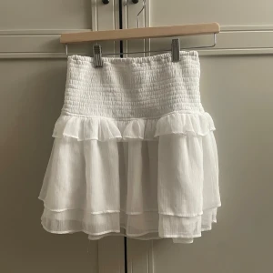 Vit volangkjol med resårmidja - Supersöt vit kortkjol med dubbla volanglager och smockad resårmidja. Kjolen är i storlek 148-164 men sitter som en xs. Använd 1-2 gånger💕