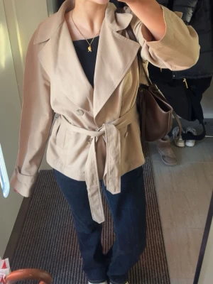 Beige trenchcoat med knytskärp - Stilren beige trenchcoat med dubbelknäppning och brett slag. Jackan har ett matchande knytskärp i midjan och är tillverkad i ett lätt tyg, perfekt för lager på lager. Klassisk och tidlös design som passar till många olika outfits.