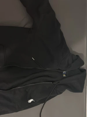 Svart hoodie från Polo Ralph Lauren - Snygg svart hoodie från Polo Ralph Lauren med dragkedja, huva med dragsko och den klassiska broderade loggan på bröstet. Tillverkad i mjukt bomullsmaterial som känns skönt mot huden. Perfekt för en avslappnad och stilren look.