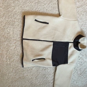 Beige teddyjacka med svarta detaljer - Mysig beige teddyjacka med svarta detaljer och dragkedja framtill. Jackan har två snedställda fickor med svarta dragkedjor och en svart bröstficka i nylon. Hög krage och avslappnad passform, perfekt för kalla dagar.Strl 158-164. Endast använd fåtal gånger. OBS: två små fläckar på baksidan, se bild. 