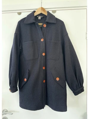 Marinblå kappa i 100% ull, H&M - Superfin mörkblå / navy / svartblå kappa / jacka / vårjacka / vårkappa från H&M i 100% ull. Det står storlek 34, men är en modell som passar alla från XS-M beroende på hur figurnära eller oversize man vill ha det. Vackra stora strukturerade bruna knappar framtill och vid ärmslut. Klassisk modell med krage och två fickor framtill i brösthöjd samt två snedställda fickor längre ner. Ärmarna är långa med en lätt puffig effekt vid axlarna och är samlade vid handleden med en knapp. Mycket gott skick!