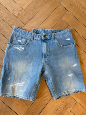 Dondup shorts - Säljer ett par blå denimshorts från Dondup i regular fit. Shortsen har slitna detaljer, fem fickor och klassisk knappgylf. Materialet är en mix av bomull och polyester, vilket gör dem både bekväma och slitstarka. Perfekta för sommaren!