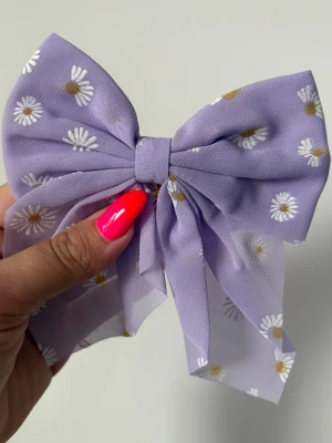 Ljuslila hårrosett med prästkragar - Söt hårrosett i ljuslila tyg med vita prästkragar och gula detaljer. Rosetten är gjord i ett tunt, lätt material och har en metallklämma på baksidan för enkel fastsättning i håret. Perfekt accessoar för att piffa upp frisyren med en blommig touch.