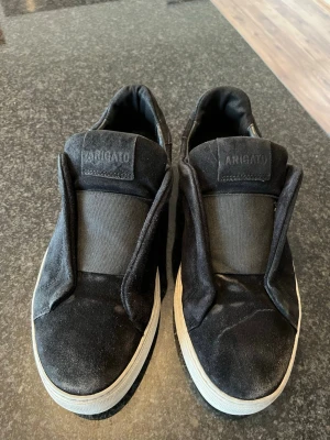 Svarta sneakers från Axel Arigato - Snygga svarta sneakers från Axel Arigato i mocka med elastiskt band över vristen och vit sula. Minimalistisk design utan snörning och diskret logga på plösen. Perfekta för dig som gillar stilrena och moderna skor. Strl 41/42 