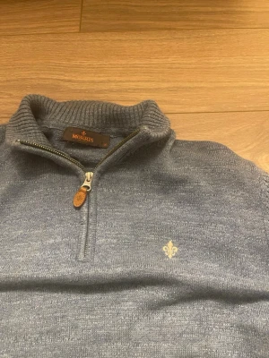 Ljusblå stickad halvzip-tröja Morris - Pris går att diskutera på en snygg ljusblå  stickad tröja från Morris med halv dragkedja och ribbade muddar. Tröjan har en liten broderad logga på bröstet och detaljer i läder vid dragkedjan. Perfekt för lager-på-lager och stilrena outfits.