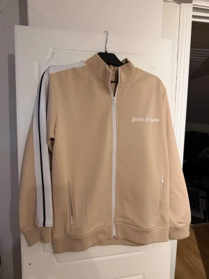  Palm Angels kofta  - Snygg beige hoodie från Palm Angels med vit dragkedja och vita samt svarta ränder längs ärmarna. Modellen har hög krage, två fickor med dragkedja och ribbade muddar. Tillverkad i ett mjukt och stretchigt material som känns sportigt och trendigt.