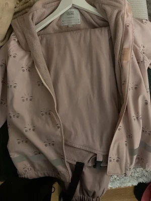 Ljusrosa jacka med kattmönster Mywear - Söt ljusrosa jacka från Mywear Young med små kattansikten som mönster. Jackan har dragkedja framtill, huva och mjukt fleecefoder på insidan. Perfekt för kyligare dagar och har reflexdetaljer vid ärmsluten.