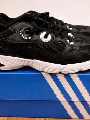 Svarta Adidas sneakers med vita detaljer - Snygga svarta Adidas sneakers med vita detaljer och logga på plösen. Skorna har en chunky vit sula, svarta meshpaneler och svarta läderdetaljer. Klassisk snörning och retroinspirerad design som sticker ut.