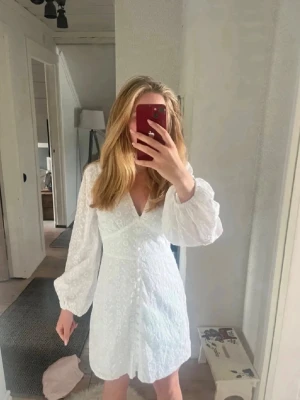 Vit kort klänning med puffärm - Söt vit kort klänning med v-ringning och puffiga långärmar. Klänningen har broderade detaljer och markerad midja som ger en feminin siluett. Perfekt för sommarens alla tillfällen.