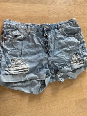 Ljusa denimshorts med slitningar - Säljer ett par ljusblå denimshorts med slitna sopade detaljer framtill och upprullade benslut. Klassisk femficksmodell med knappgylf och avslappnad passform. Perfekta för var dodg sommaroutfit!