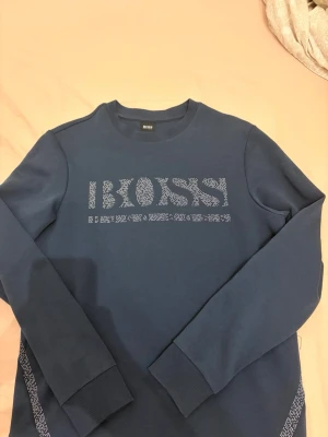 Blå sweatshirt från BOSS - Snygg blå sweatshirt från BOSS med rund halsringning och diskret logotyp i mönstrad stil på bröstet. Tröjan har långa ärmar och ribbade muddar vid ärmslut och nederkant. Perfekt för en avslappnad och trendig look.