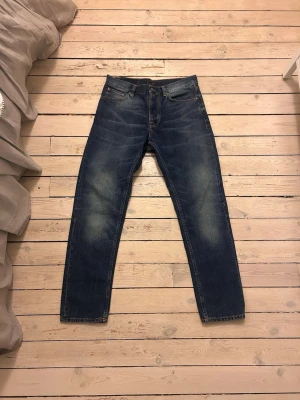 Blå jeans från Nudie Jeans Co - Säljer ett par klassiska blå jeans från Nudie Jeans Co med raka ben och snygga slitningar. Jeansen har orange kontrastsömmar, fem fickor och är tillverkade i denim. Perfekta för en avslappnad och stilren look.