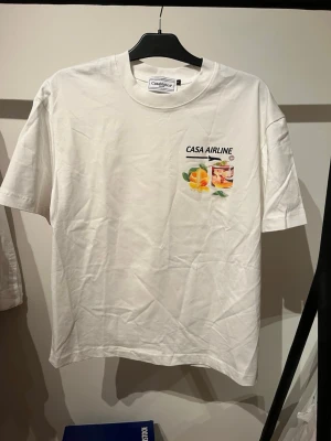 Vit CASA AIRLINE t-shirt med tryck Oversized  - Vit t-shirt från Casa Blanca med färgglatt tryck av drink och apelsiner på bröstet samt stort motiv på ryggen med flygplansfönster, solnedgång och texten CASA AIRLINE. Klassisk passform och rund hals. Perfekt för dig som gillar unika prints.SITTER SOM EN M ELLER S OVERSIZED 