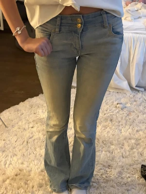 Ljusblå bootcut jeans med dubbla knappar - Säljer ett par ljusblå bootcut jeans med dubbla guldknappar i midjan och klassiska fem fickor. Jeansen har en lätt utsvängd passform nedtill och är tillverkade i denim. Perfekta för en avslappnad och trendig look.