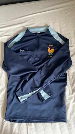 Nike blå FFF träningströja - Blå långärmad träningströja från Nike med FFF-logga och tupp på bröstet. Tröjan har ljusblå detaljer på axlar och ärmar samt halv dragkedja framtill. Perfekt för fotbollsträning eller sportiga aktiviteter.
