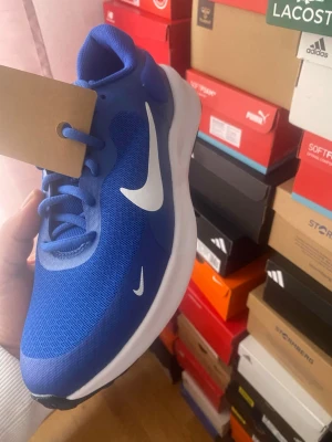 Blå Nike sneakers med vit sula - Snygga blå sneakers från Nike med vit sula och stora vita swoosh-loggor på sidan. Ovandelen är i meshmaterial för bra andningsförmåga och skon har klassisk snörning. Perfekt för dig som gillar sportig och stilren look.
