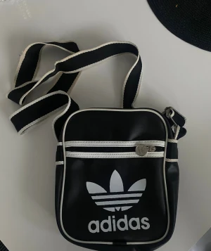 Svart axelväska från Adidas - Snygg svart axelväska från Adidas med vit logga och detaljer. Väskan har ett stort fack och en mindre ficka med dragkedja framtill. Justerbar axelrem i svart med vit kant. Materialet är syntet och insidan är fodrad. Perfekt storlek för mobil, plånbok och nycklar.