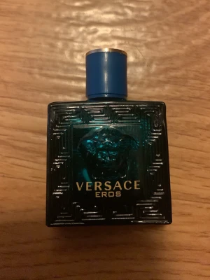 Versace Eros Eau de Toilette - Versace Eros parfym 5ml i en stilren, fyrkantig flaska av mörk turkost glas med guldtext och Medusa-huvud på framsidan. Flaskan har ett blått lock med gulddetalj och ett elegant, grekiskt mönster i glaset. En ikonisk doft för dig som gillar exklusiva parfymer.