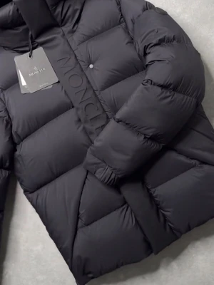 Svart dunjacka från Moncler - Snygg svart dunjacka från Moncler med quiltad design och stora puffiga partier. Jackan har tryckknappar och en bred panel med Moncler-logga längs dragkedjan. Perfekt för kalla vinterdagar och riktigt trendig look.