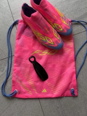 Adidas F50 rosa fotbollsskor - Säljer ett par Adidas F50 fotbollsskor i en riktigt snygg rosa färg med gula och blå detaljer. Skorna har strömlinjeformad design, låg vikt och greppvänlig dobbsula i gult. Ovandelen är i syntetmaterial och de kommer med en matchande rosa påse och skohorn.
