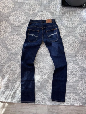 svarta originella jeans från Nudie - Säljer ett par mörkblå jeans från Nudie med klassisk rak passform. Jeansen har kontrastsömmar, två bakfickor with Nudies 'wave'-brodyr och läderpatch i midjan. Materialet är slitstark denim i bomull. Perfekta för dig som gillar stilrena och tidlö gor jeans.
