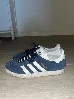 Adidas Gazelle blå sneakers -  Adidas Gazelle sneakers i blå mocka med vita detaljer och klassiska tre ränder på sidan. Storlrk 40, mycker kvar att ge!