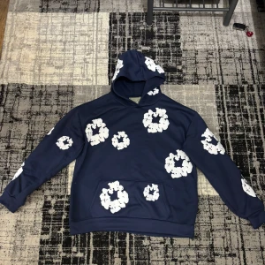  hoodie från Denim Tears - Säljer en mörkblå hoodie från Denim Tears med stora vita blommönster över hela tröjan. Hood fork fr hourly och magficka framtid. Perfekt för dig som vill stick PN ut med en unik och trendig stil. Priset kan diskuteras vid snabb affär. 
