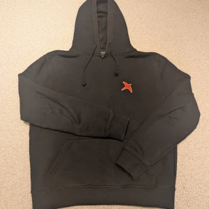 Svart hoodie med röd fågelpatch - Svart hoodie med huva och dragsnören samt en röd fågelpatch broderad på bröstet. Klassisk känguruficka framtill och ribbade muddar vid ärmslut och nederkant. Perfekt för en chill och stilren look.