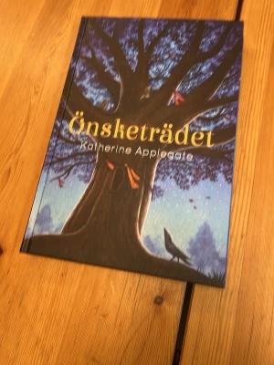 Önsketrädet av Katherine Applegate - En magisk och stämningsfull bok med hårda pärmarA, 'Önsketrädet' av Katherine Applegate. Omslaget har en stor trädillustration i blå och bruna toner med guldfärgad text. Perfekt för dig som gillar berättelser om vänskap, hopp och natur.