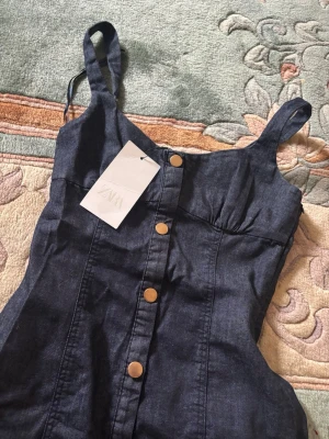 Mörkblå jeansklänning från Zara - Mörkblå långklänning från Zara i jeansmaterial med axelband och knappar framtill. Klänningen har en figurnära passform och markerad midja, perfekt för dig som gillar en stilren och trendig look.