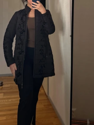 MICHA jacka - Svart jacka med ett detaljerat mönster. Jackan har en dragkedja framtill och två fickor med dragkedja. Insidan av jackan har ett färgglatt blommigt mönster med fåglar och fjärilar.