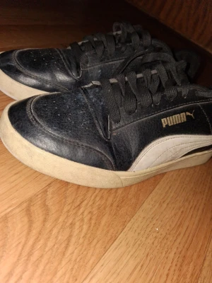 Svarta Puma sneakers med vit sula - Snygga svarta sneakers från Puma med klassisk vit sula och vit sidodetalj. Skorna har svarta skosnören och är tillverkade i läderliknande material. Perfekta för en sportig och stilren look.
