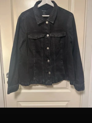Svart jeansjacka från Lindex - Snygg svart figursydd jeansjacka från Lindex med klassisk krage, silverfärgade knappar och två bröstfickor med lock. Jackan har raka ärmar och markerade sömmar för en tidlös look. Perfekt att slänga över en hoodie eller t-shirt.