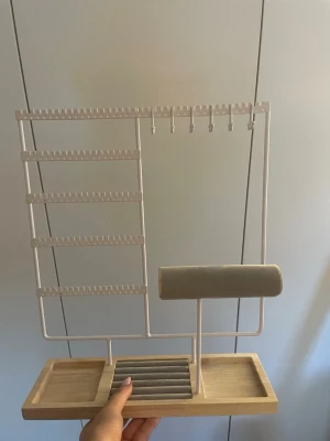 Smyckesställ i trä och metall - Snyggt smyckesställ i ljust trä och vit metall med flera rader för örhängen, krokar för halsband och en vadderad rulle för ringar eller armband. Perfekt för att organisera dina smycken på ett stilrent sätt i rummet.