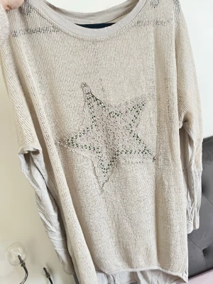 Beige stickad tröja med stjärna - Beige stickad tröja med avslappnad passform och rund halsringning. Framtill finns en stor stjärna i spets och små pärlor som detalj. Tröjan har långa ärmar och är tillverkad i ett tunt, mjukt material som ger en luftig känsla.