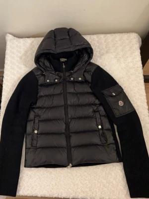 Svart cardigan  från Moncler - Snygg svart cardigan från Moncler med stickade ärmar och huva. Jackan har dragkedja framtill, två fickor med tryckknappar och en ikonisk Moncler-logga på ärmen. Perfekt för kyliga dagar och riktigt trendig look.